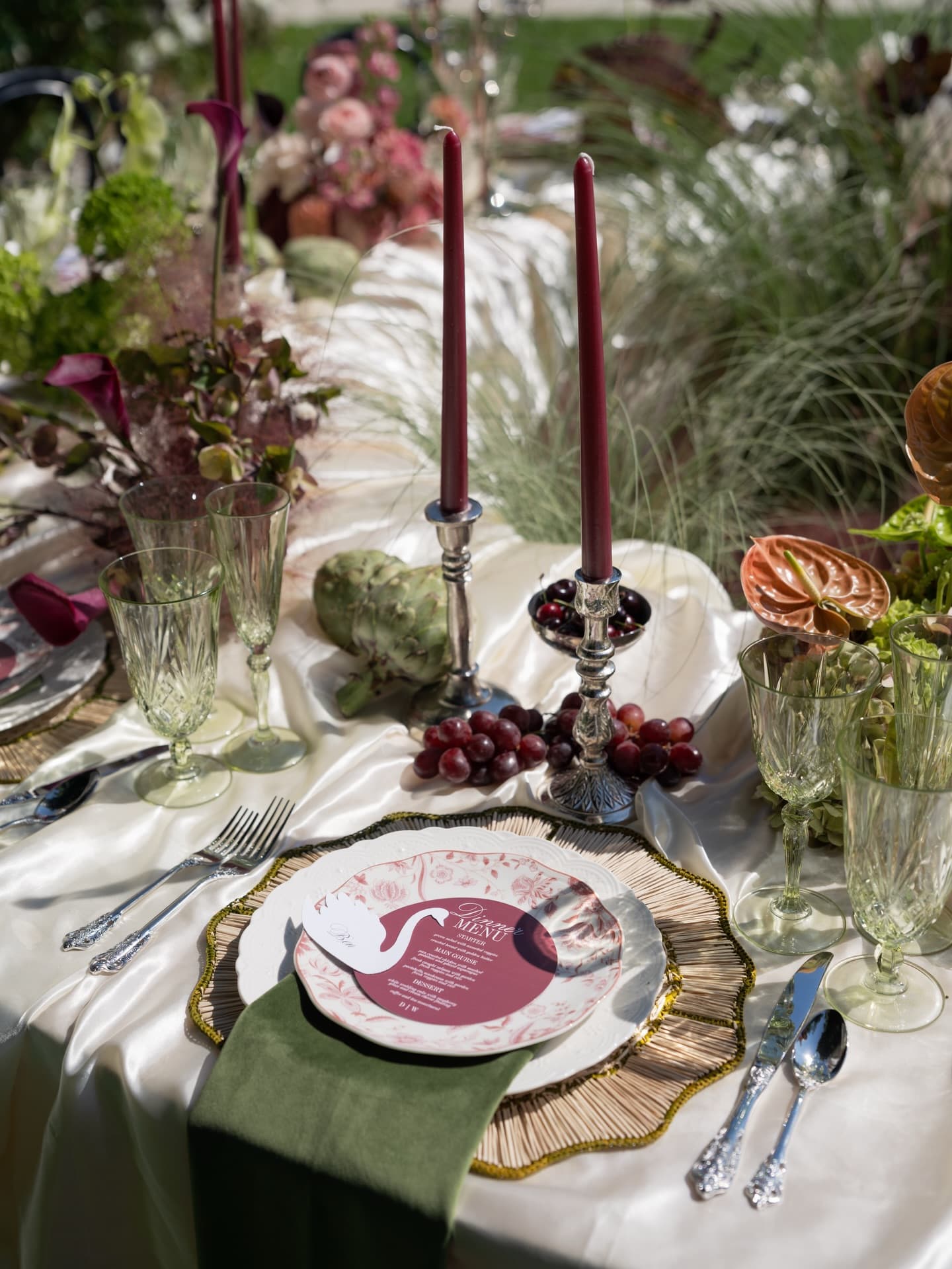A place setting with a swan-printed menu on a gold-rimmed charger, an olive velvet runner, silver flatware and dark florals