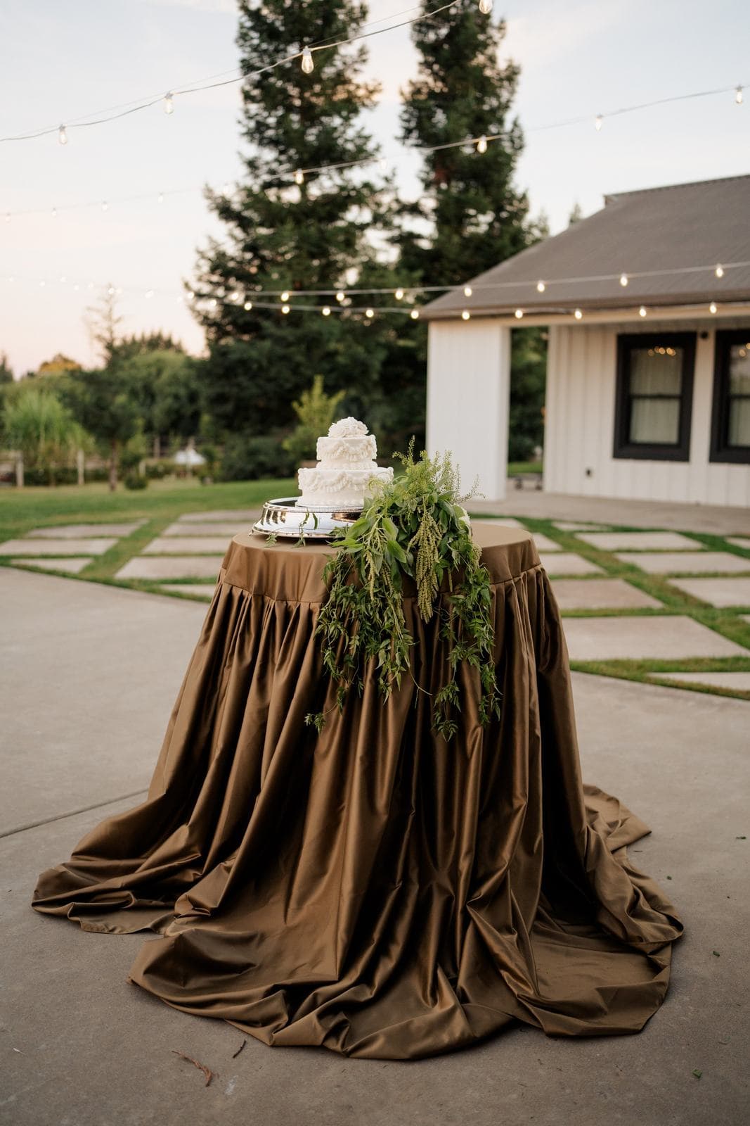 A table draped to the floor in olive-bronze linen beneath string lights at dusk