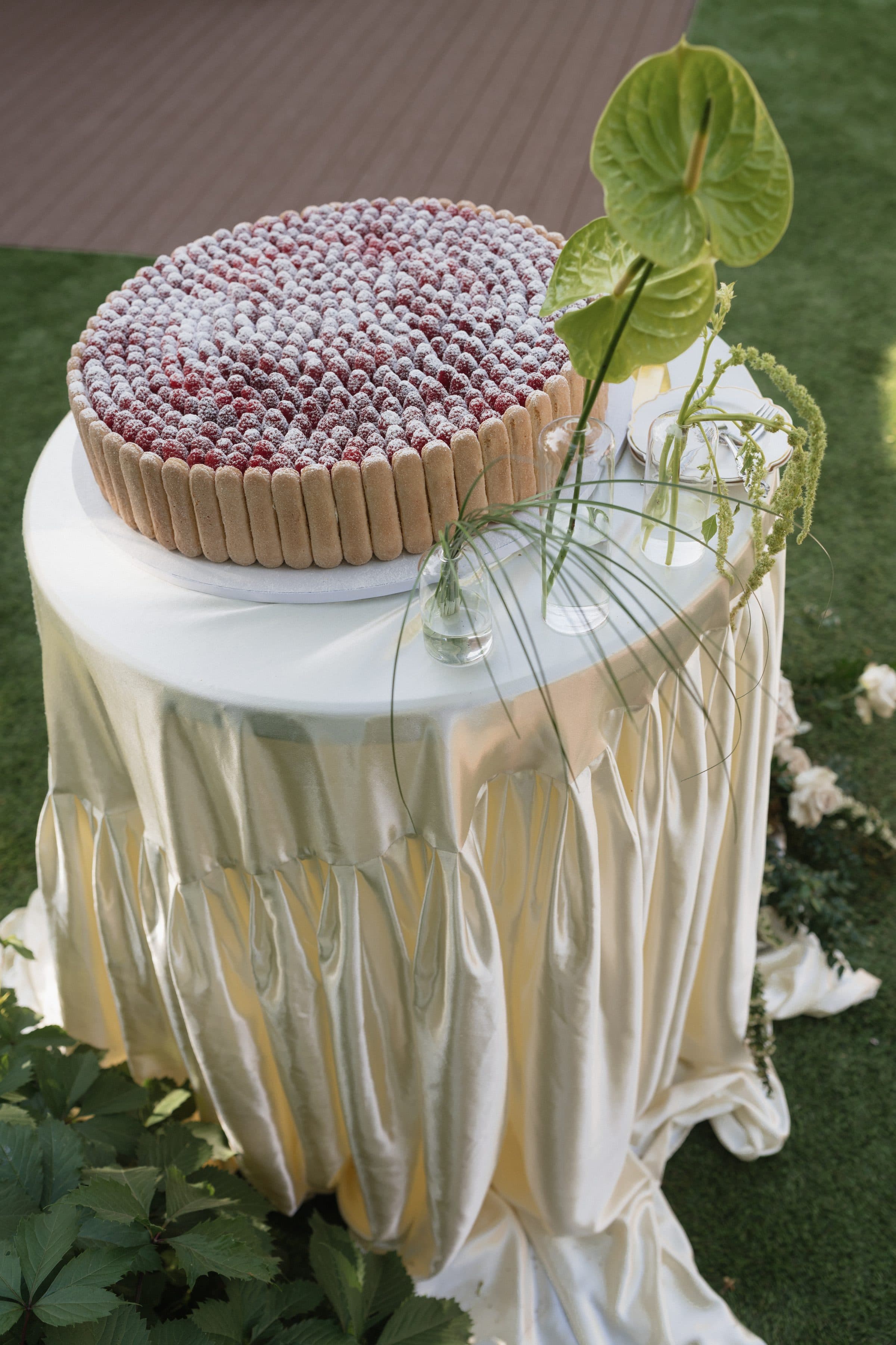 A dessert table skirted in gathered ivory satin, a raspberry tart on top and a green anthurium beside it, on a lawn