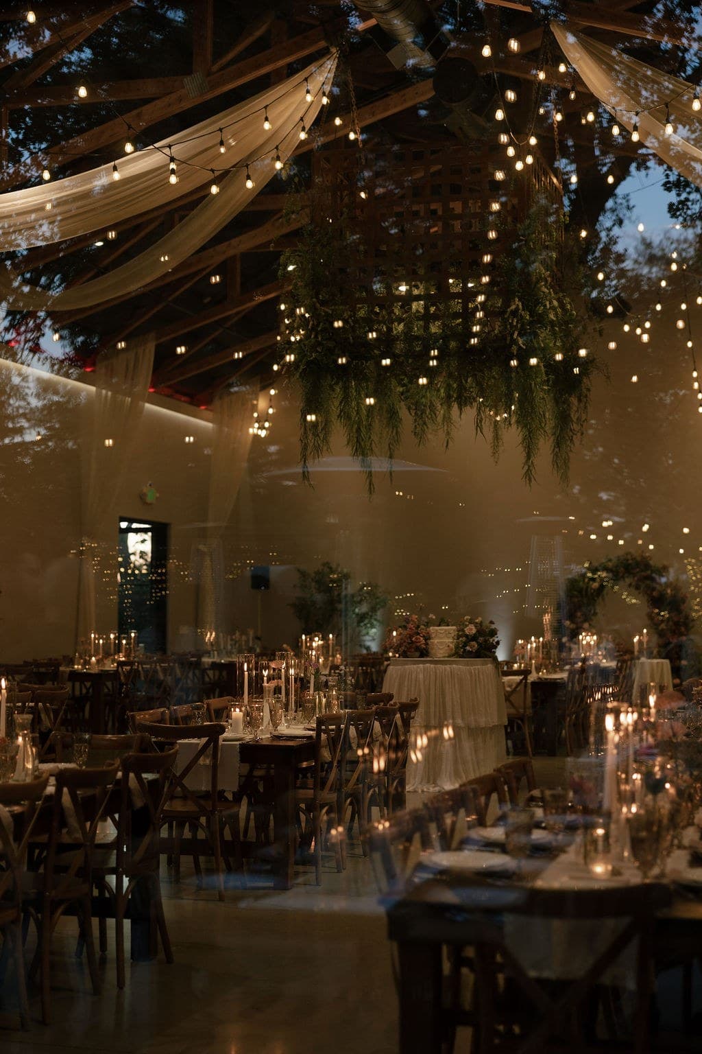 A candlelit evening dinner beneath string lights and suspended greenery, farm tables set with tapers and crystal in a rustic pavilion