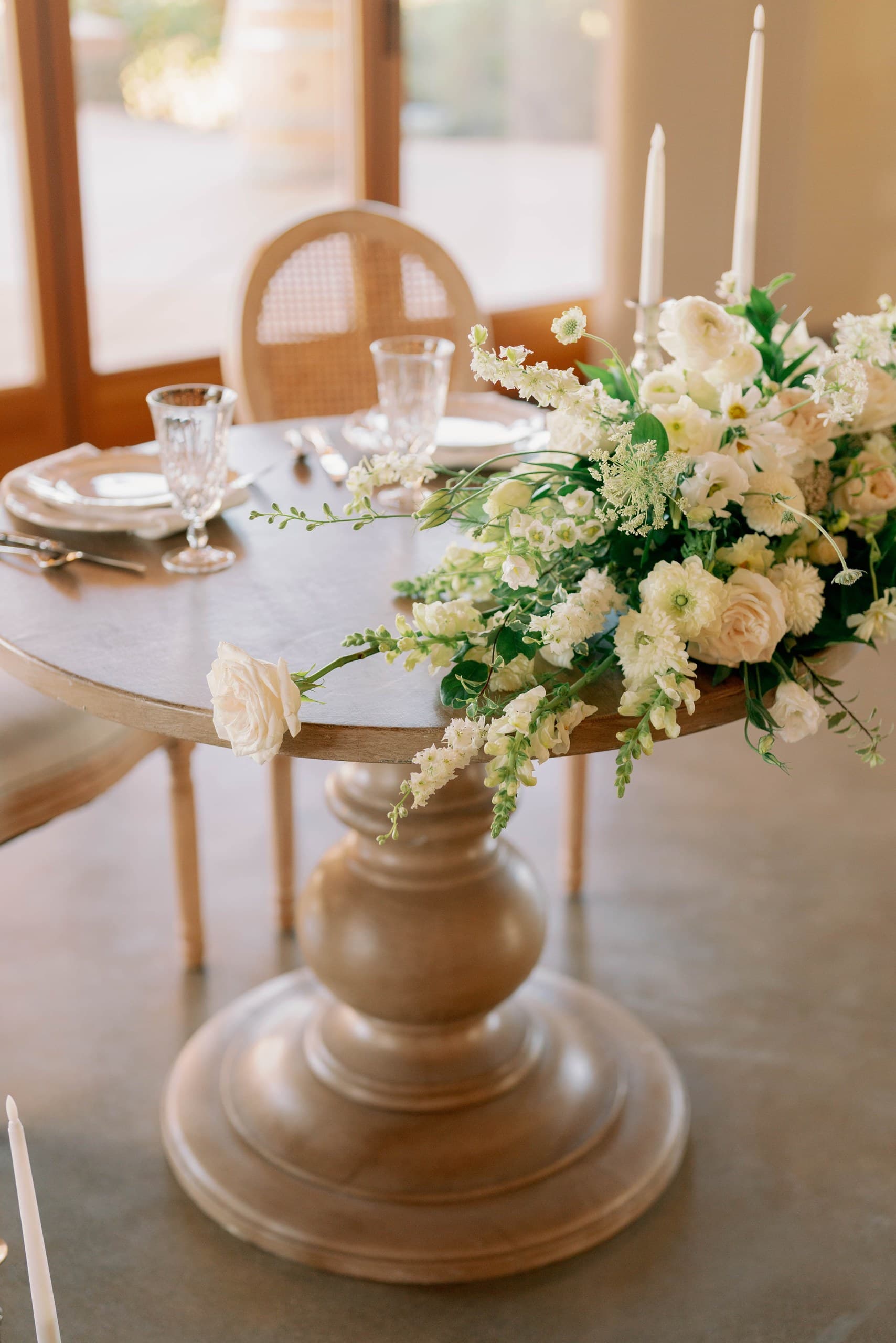 A round wooden table set with a low white-and-green centerpiece, taper candles, wine glasses and cane-back chairs