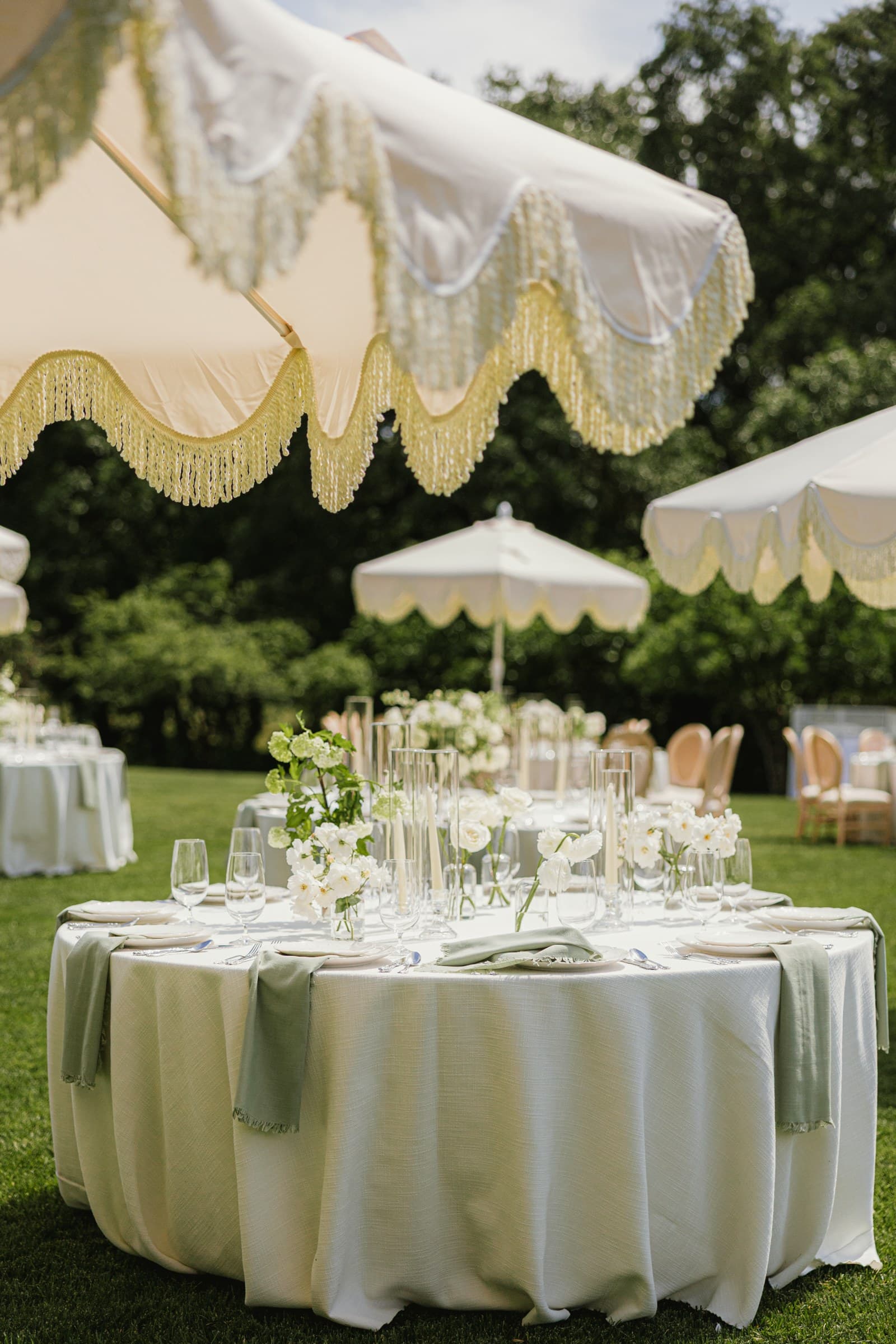 Sage-green banquet tables outdoors beneath scalloped fringed umbrellas, low white florals and champagne linens on the lawn