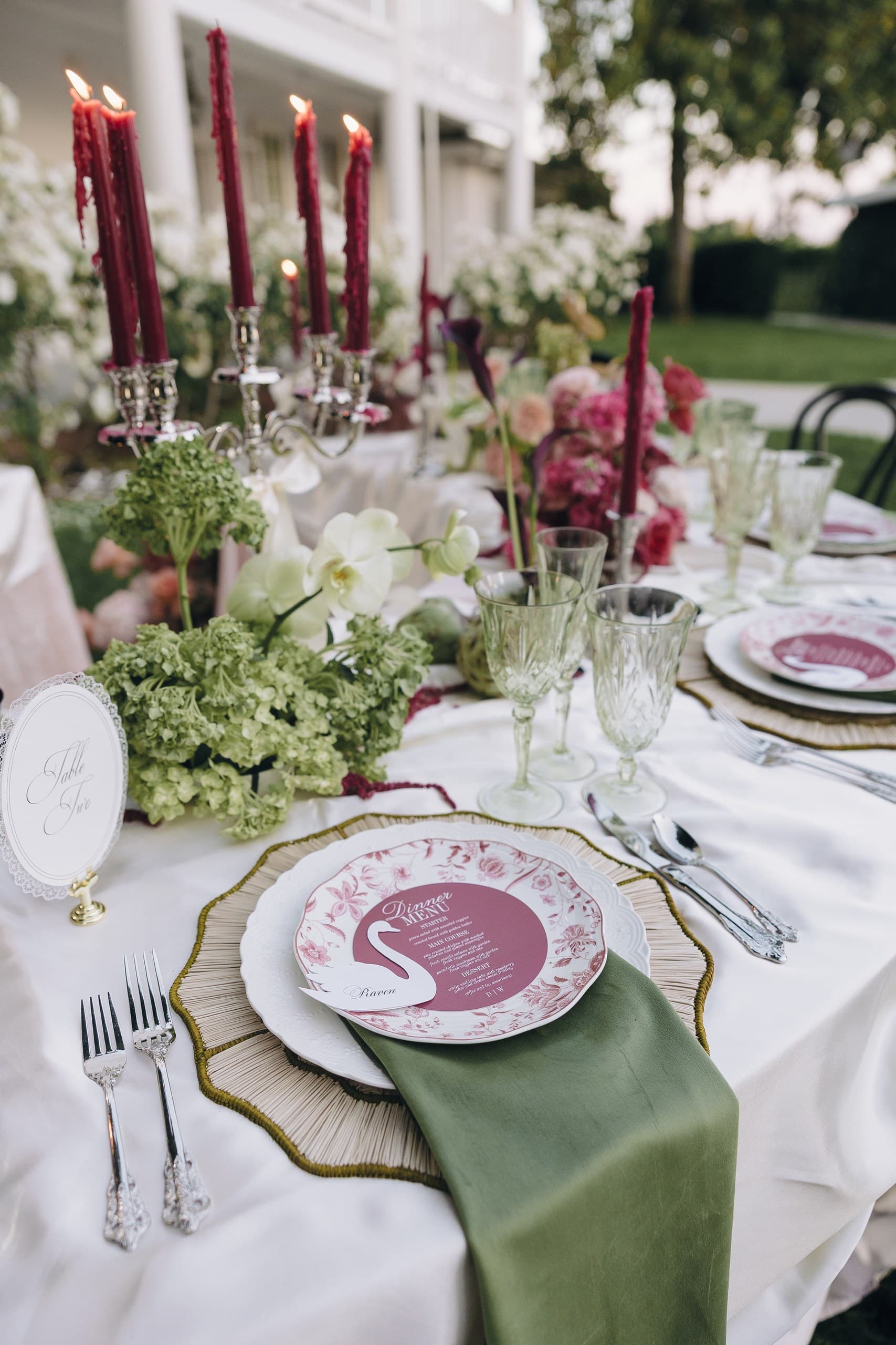 A place setting with a swan-printed menu on a scalloped charger, burgundy tapers in a silver candelabra, an olive napkin and pink florals