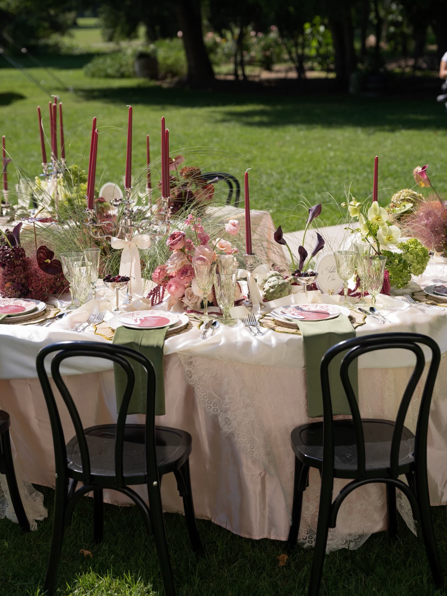 A dinner table set with burgundy florals, dark candles and a velvet runner in full garden daylight