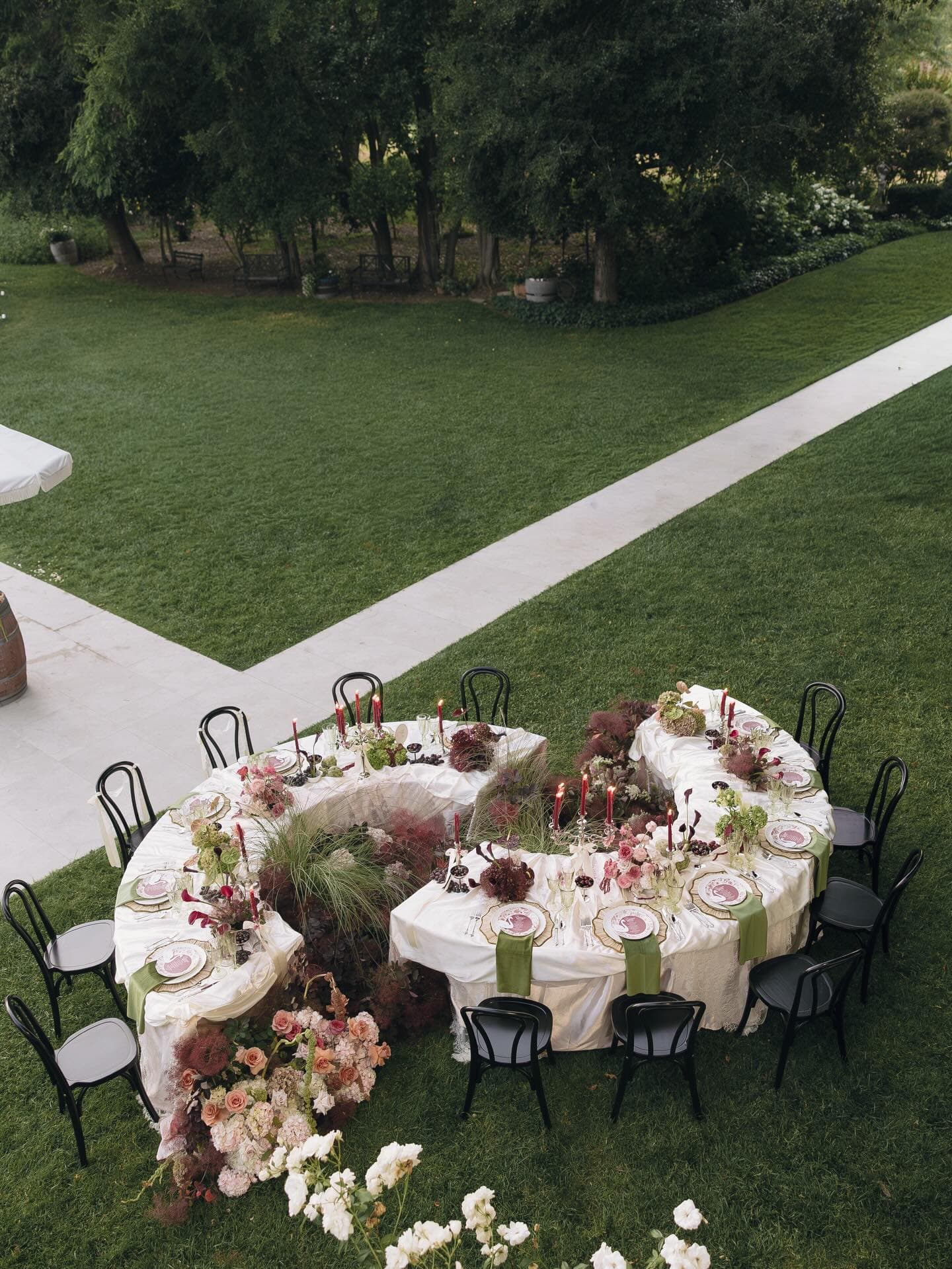 A long, curved dinner table set on a lawn beside a white house, seen from above with chairs all around