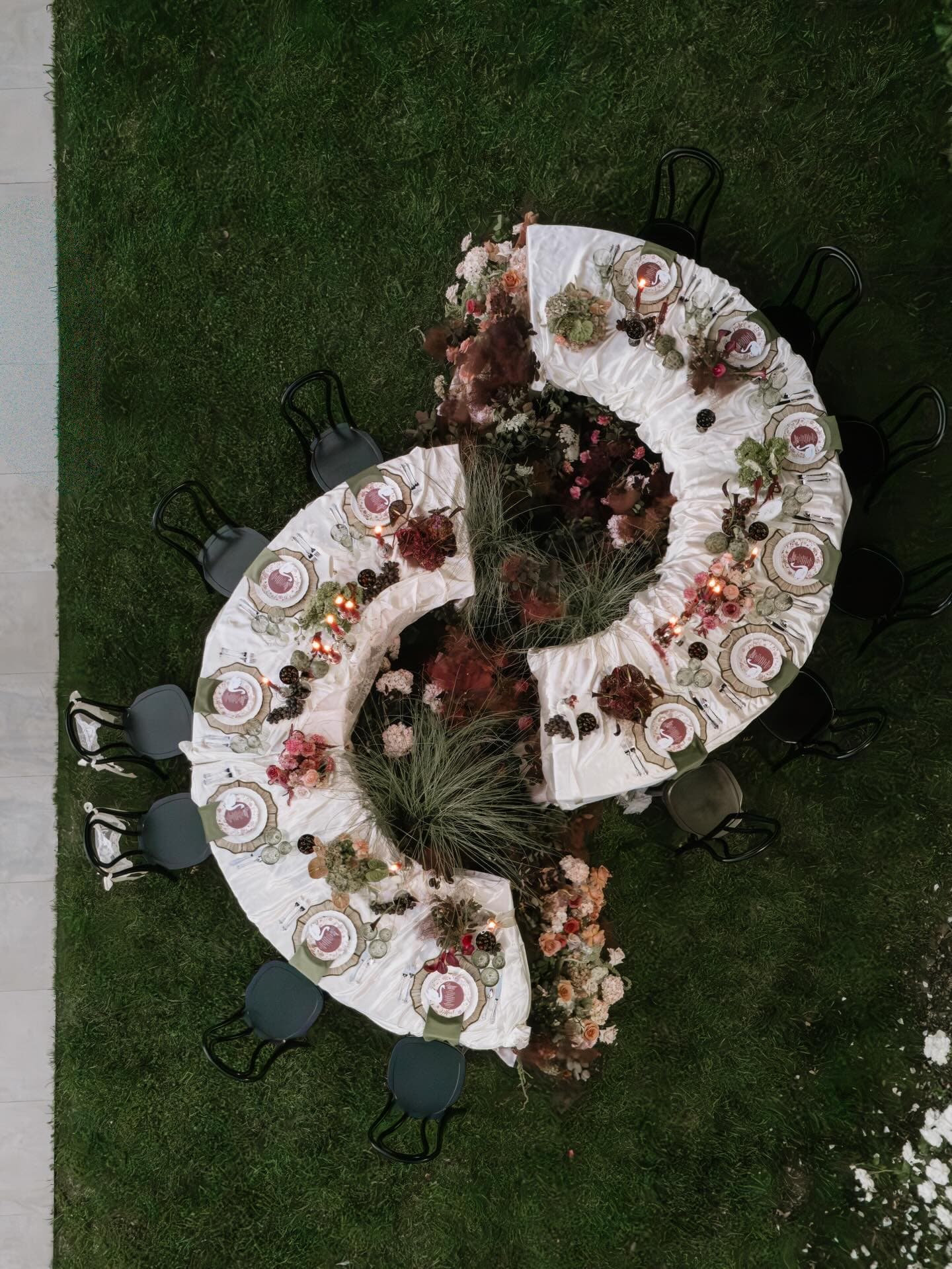 A serpentine dinner table set with florals and candles, seen from above beside a vineyard