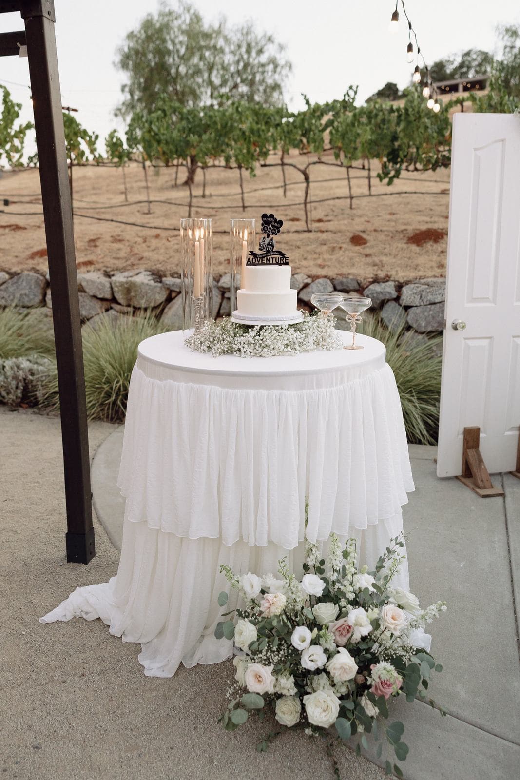 A tiered white linen table set with a cake and flowers in a vineyard