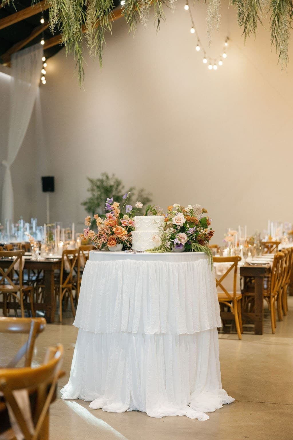 A two-tier white cake on a full-length ivory ruffle-draped table, flanked by peach-and-lilac florals, farm tables and cross-back chairs waiting behind under string lights