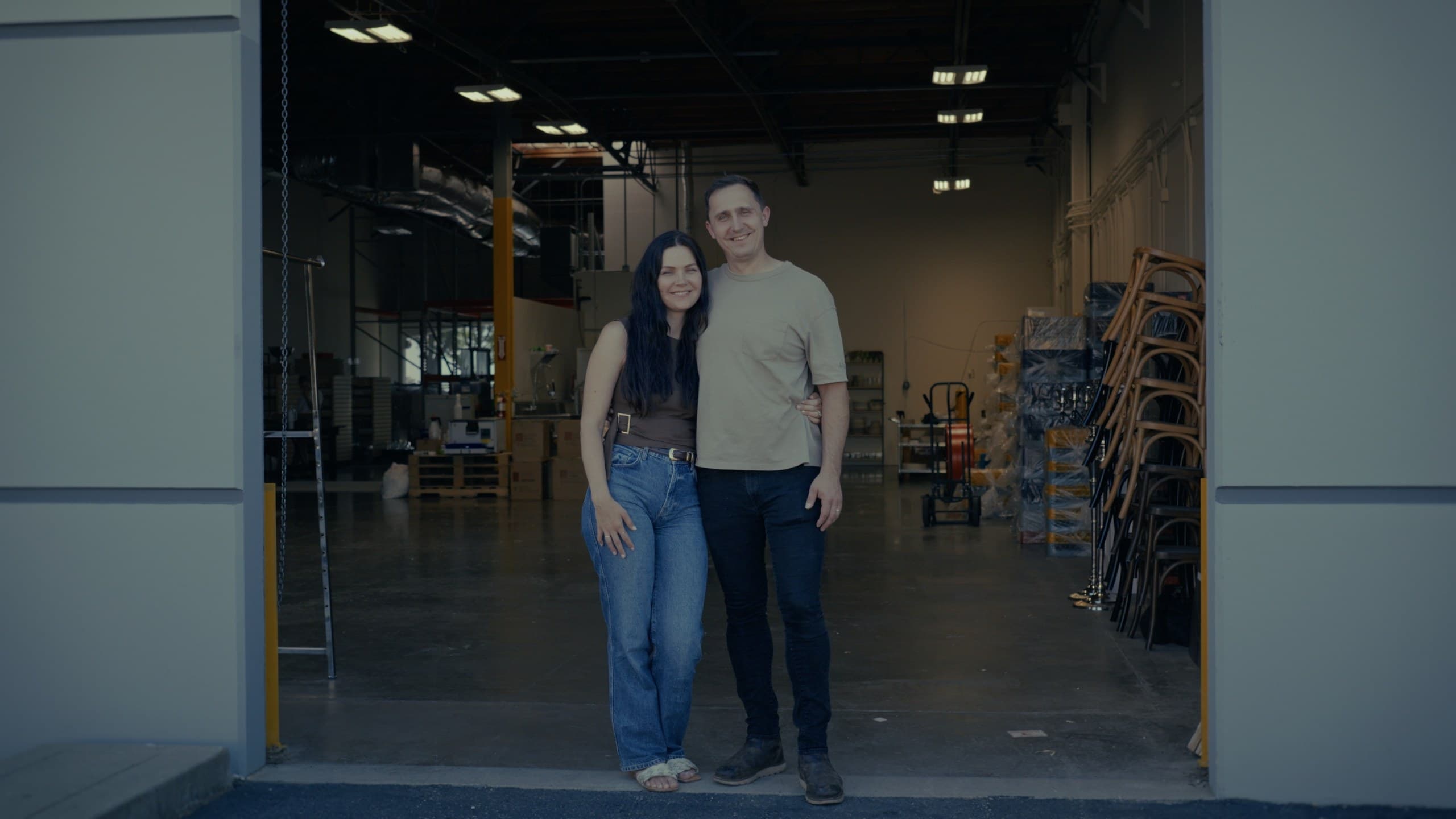 The founders standing in the open roll-up door of the Lincoln warehouse, folding chairs and event inventory racked behind them