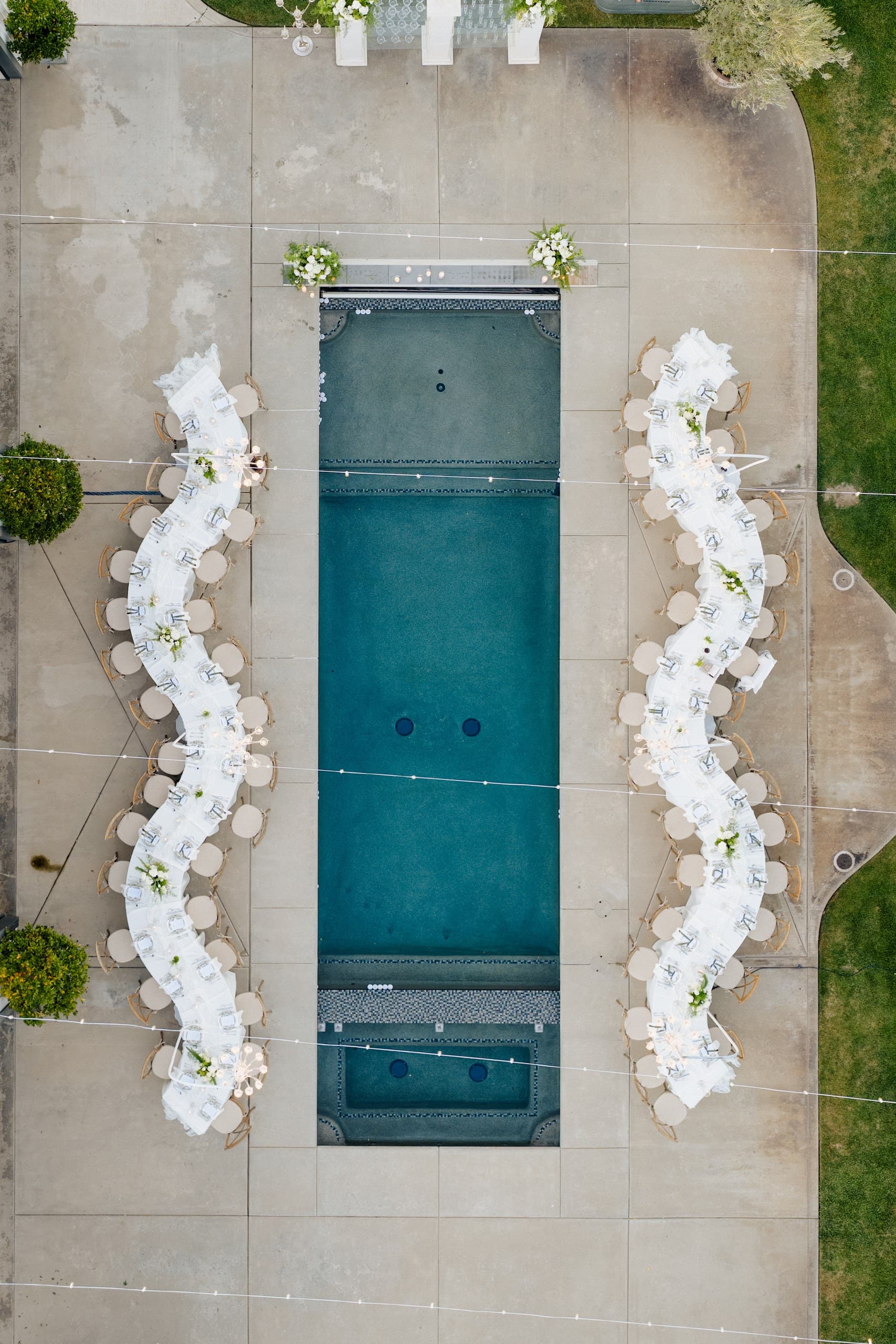 Two long white banquet tables curving along either side of a rectangular pool, seen from above with a rehearsal-dinner or corporate reception set out
