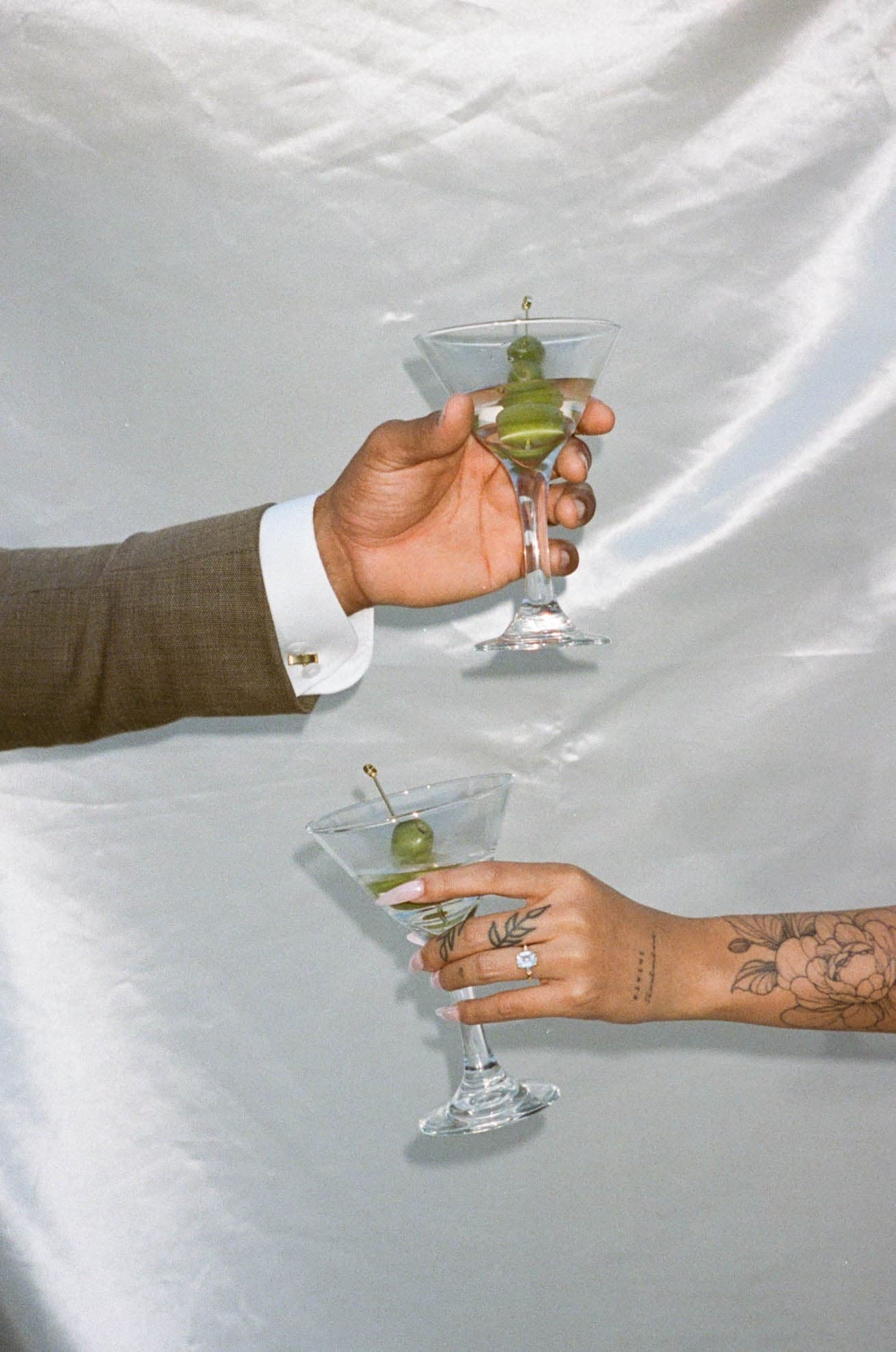 Two hands raising olive martini glasses in a toast against ivory drapery, an engagement diamond on the ring finger