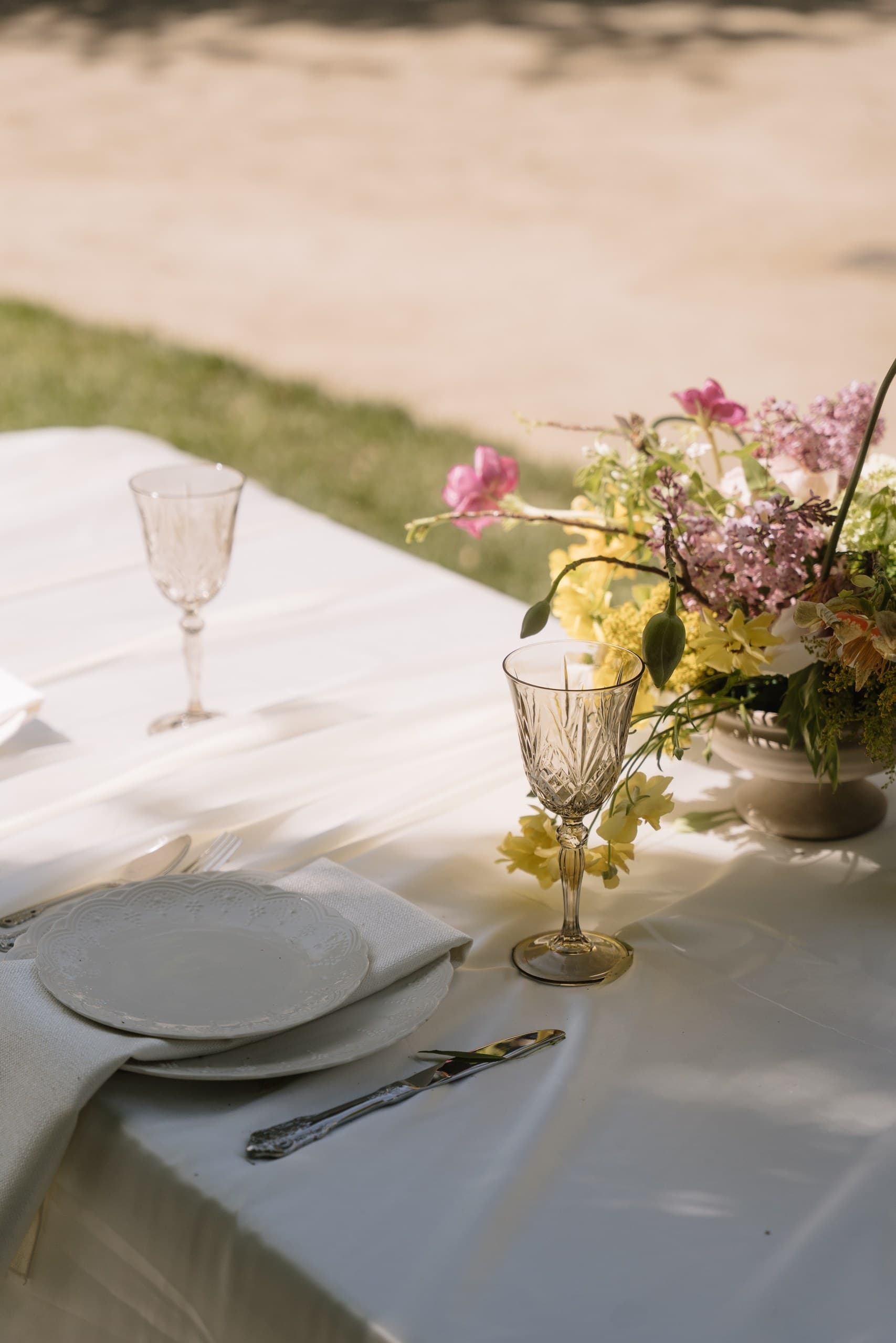 An outdoor place setting on ivory linen with a yellow-cut crystal glass and a spring bouquet of lilac, yellow and pink flowers
