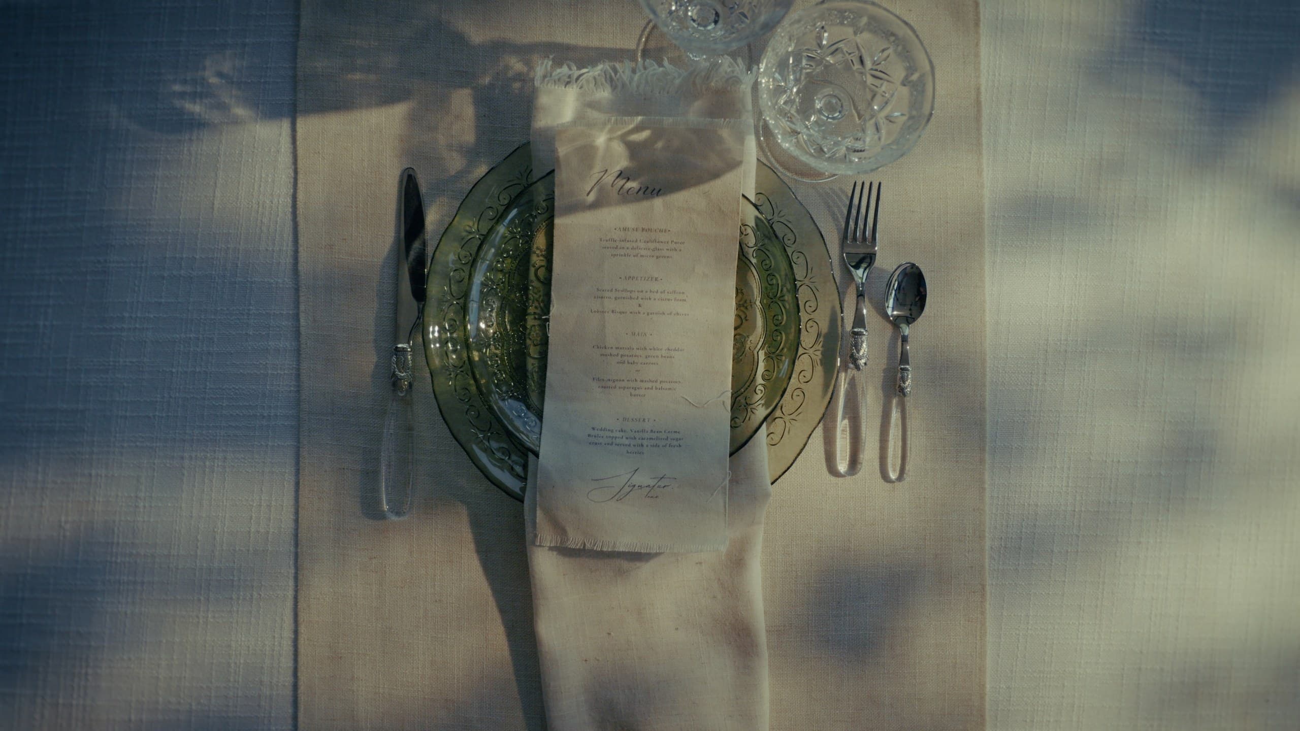 A place setting seen from above through dappled shade — a printed menu on a scalloped green plate, silver flatware, cut-crystal coupes, a linen napkin