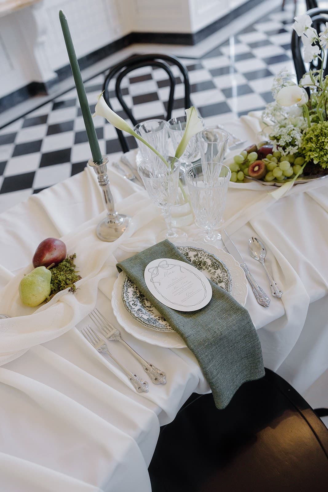 A place setting with an olive napkin, silver flatware and a green calla lily on a black-and-white checkerboard floor beneath black bentwood chairs