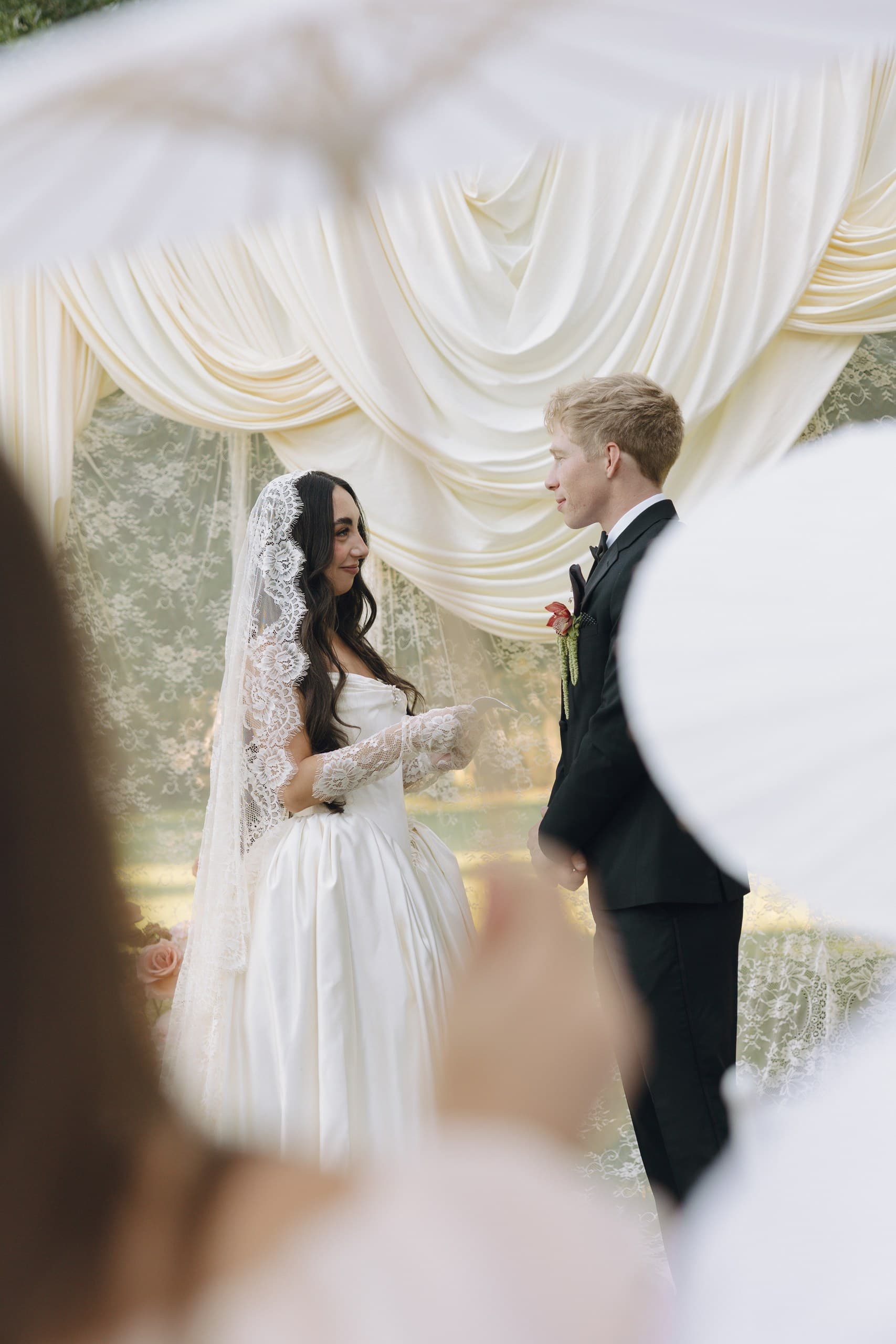 A bride in a lace veil and satin gown facing her groom beneath an ivory drapery arch, framed by a guest's paper fan in the foreground